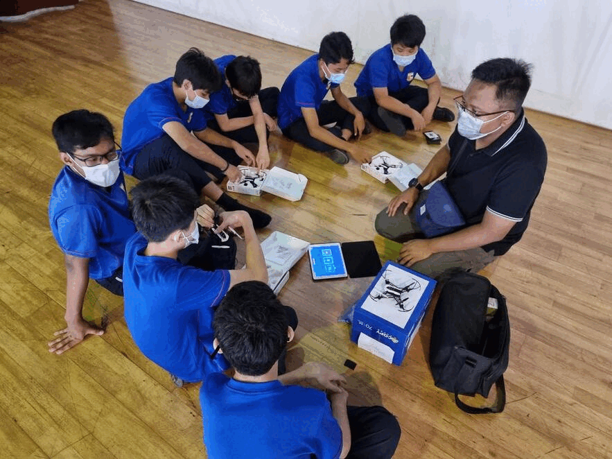 A masked adult instructor and masked students in blue shirts sit on a floor examining drone boxes and parts.